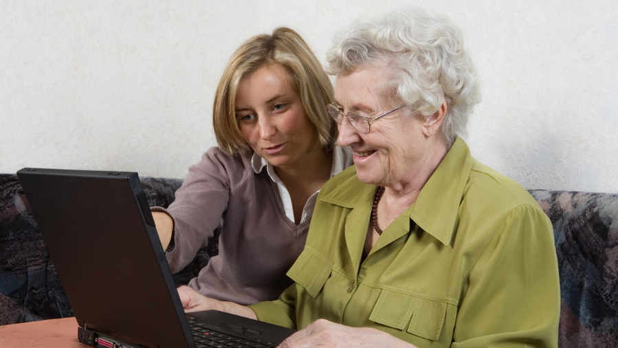 Eine ältere und eine jüngere Frau schauen in einem Laptop. Die jüngere erklärt der älteren Frau etwas.