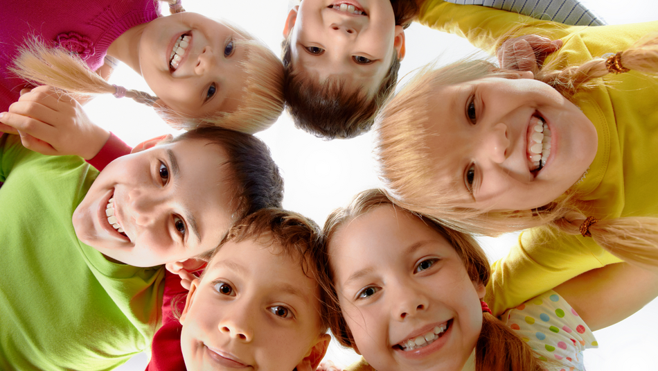 Sechs Kinder im Grundschulalter stecken in einem Kreis die Köpfe zusammen und schauen lächelnd nach unten.