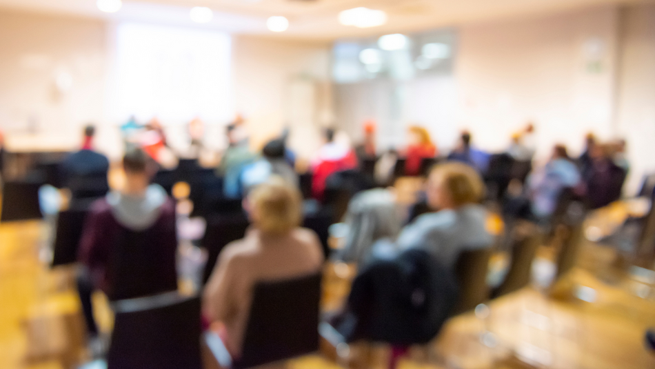 Verschwommen dargestellt: Mehrere Reihen mit Personen auf Stühlen hören einem Vortrag zu.