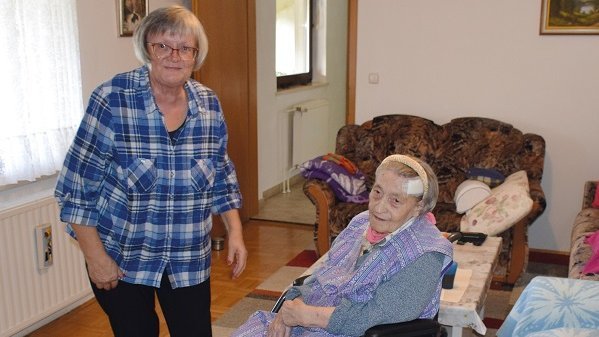 In einer Wohnung steht eine Frau neben einer älteren und sitzenden Frau im Rollstuhl. Beide schauen in die Kamera.