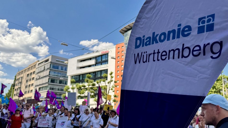 Kundgebung der Diakonie Württemberg auf dem Rotebühlplatz in Stuttgart. Zu sehen sind Mitarbeitende der Diakonie Württemberg mit Plakaten und im Vordergrund eine Beach-Flag der Diakonie Württemberg.
