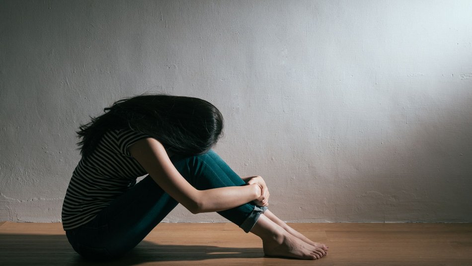 Bild zeigt eine junge Frau die mit angezogenen Beinen auf dem Boden sitzt ihr Kopf ist zwischen die Arme gesenkt