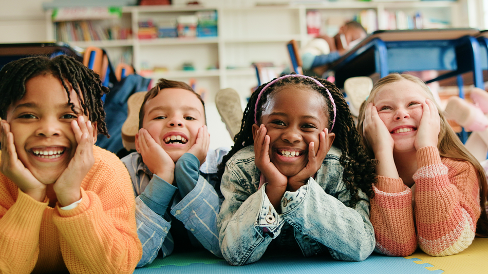 Vier lachende Kinder mit unterschiedlichen Hautfarben liegen auf dem Boden und stützen ihre Köpfe auf ihren Armen ab.