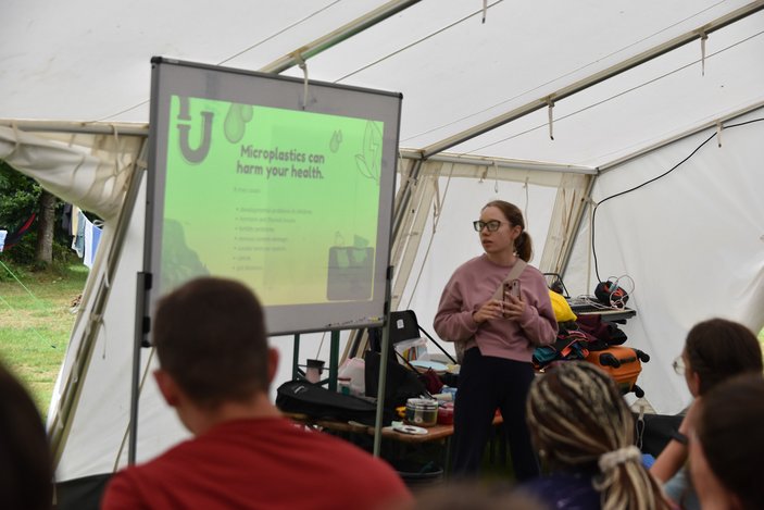 Eine Präsentation wird in einem Zelt auf eine Leinwand projiziert. Davor steht eine Frau, die in ein Publikum spricht.