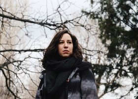 Eine Frau mit dunklen Haaren befindet sich im Freien und blickt in die Ferne.