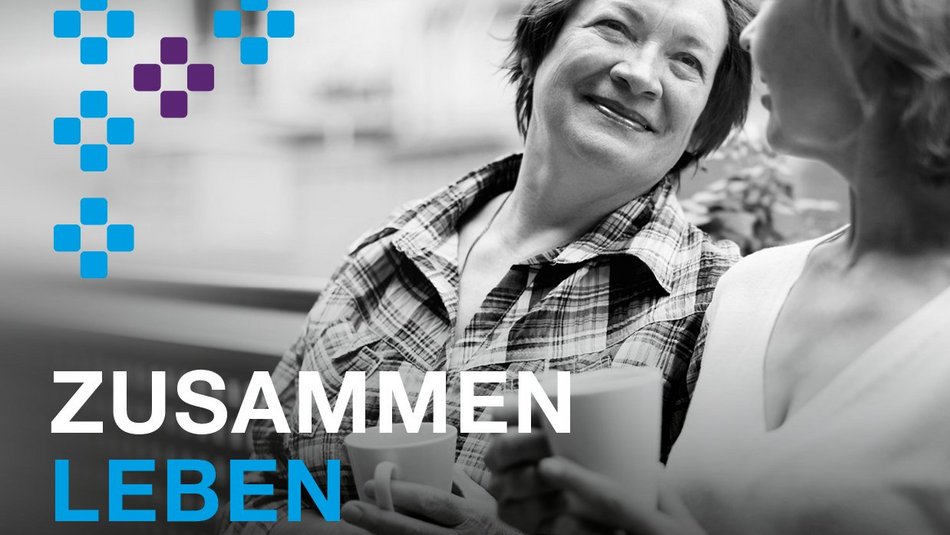Das Bild zeigt das Jahresmotto "zusammen leben": Zwei Frauen haben Kaffeetassen in der Hand und lächeln sich an