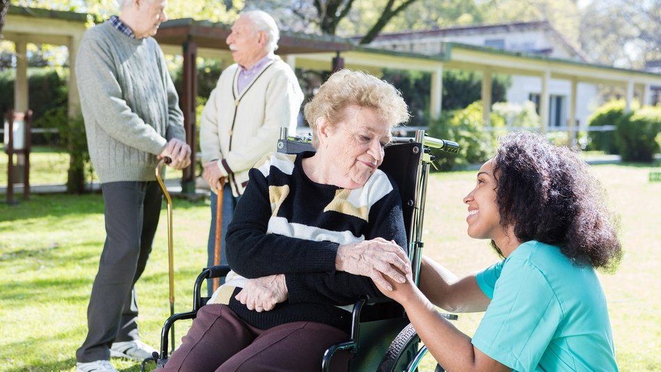 Eine Pflegerin kniet neben einer älteren Frau, die im Rollstuhl sitzt. Beide lächeln sich an. Im Hintergrund unterhalten sich zwei ältere Männer.