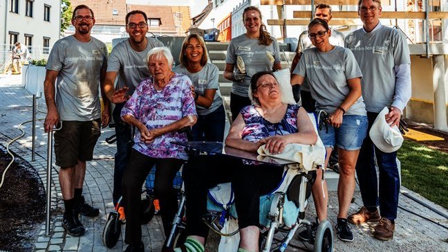 Mehrere Personen eines Unternehmens stehen hinter Menschen, die im Rollstuhl sitzen. Alle lachen in die Kamera.