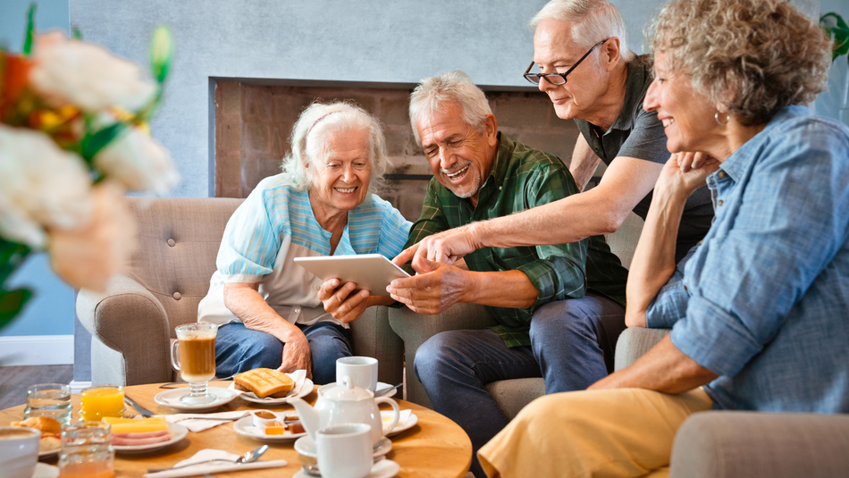 Vier ältere Menschen sitzen bei Kaffee und Kuchen an einem Tisch und schauen in ein Tablet. Sie lachen und sind fröhlich.