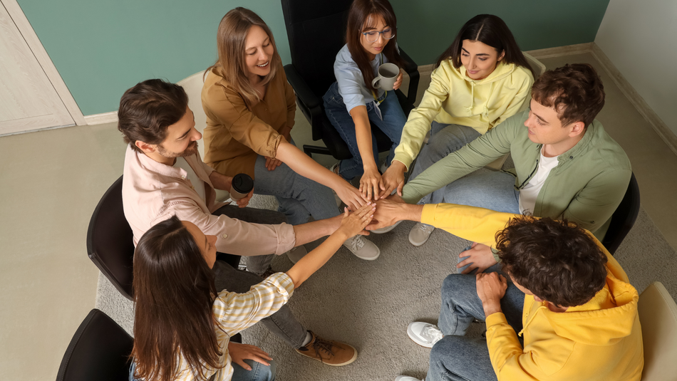 Sieben Jugendliche sitzen im Kreis auf Stühlen und halten die Hände in der Mitte des Kreises zusammen.