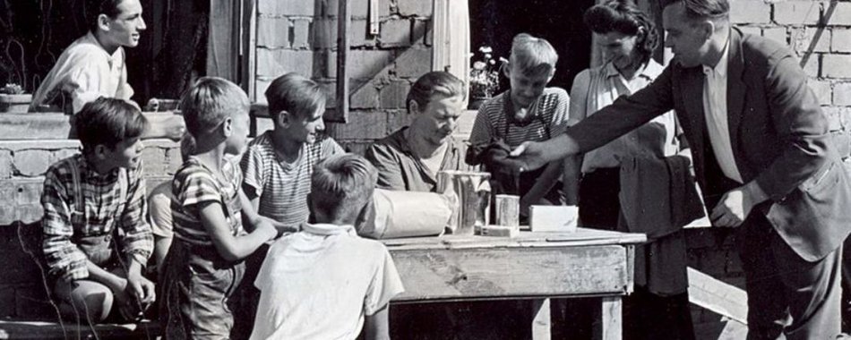 Kinder und Erwachsene sitzen in früheren Zeiten im Freien an einem Tisch.