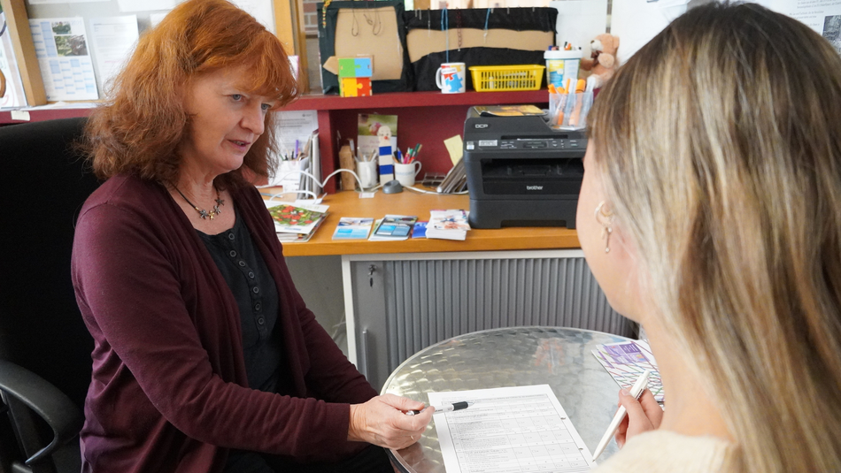 Beratungssituation zwischen einer diakonischen Beraterin und einer jungen Frau an einem runden Tisch. Die Beraterin zeigt mit einem Stift auf ein Blatt Papier.