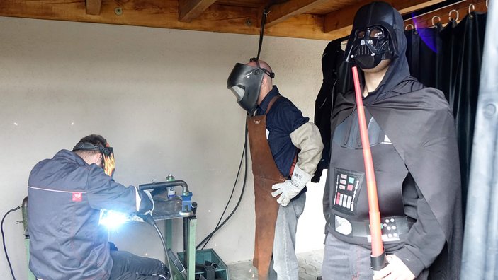 Eine Darth Vader Figur steht neben 2 Personen die schweißen. 