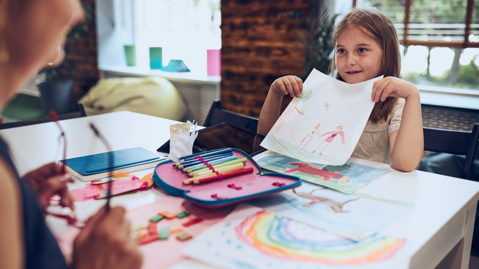 Ein Mädchen im Grundschulalter zeigt einer Erwachsenen ein selbstgemaltes Bild einer Familie.