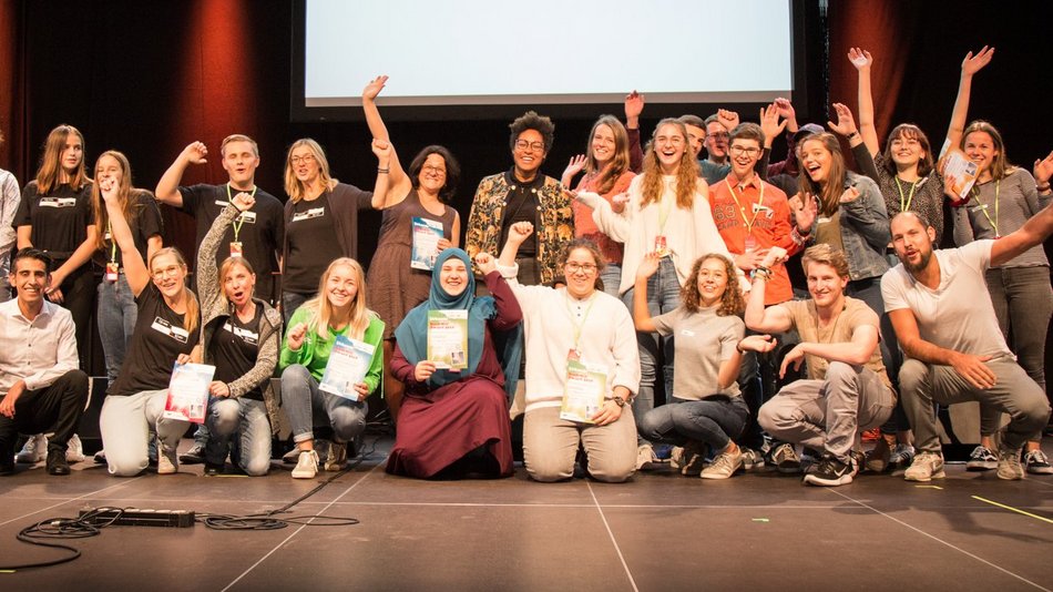 Preisträger und Preisträgerinnen des Jugenddiakoniepreis stehen auf einer großen Bühne und lachen und winken in die Kamera.