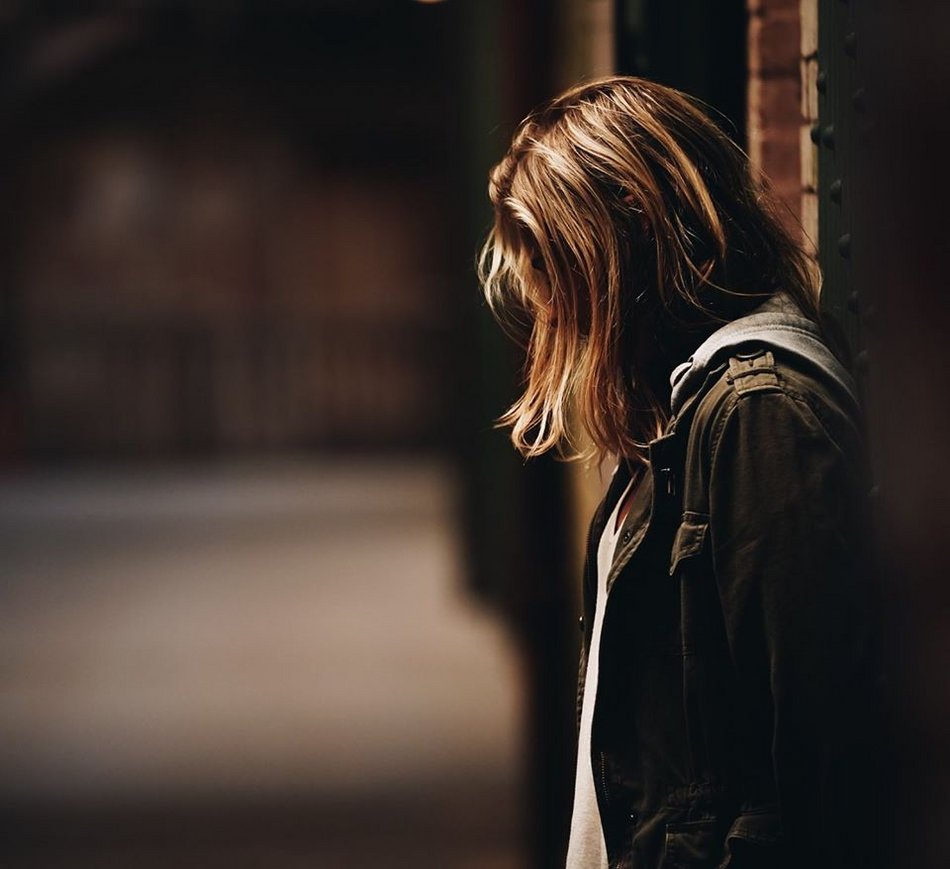 Eine junge Frau mit schulterlangen Haaren ist von der Seite zu sehen und lehnt mit dem Rücken an einer Wand. Ihr Kopf ist gesenkt und die Haare überdecken ihr Gesicht.