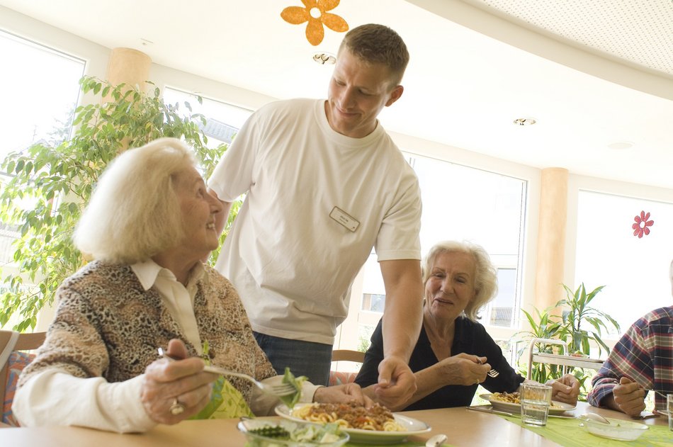 Bild zeigt einen Betreuer wie er einer älteren Frau etwas reicht, beide schauen sich an und lächeln