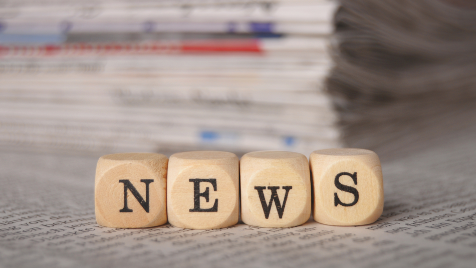Mit Würfeln ist das Wort News dargestellt. Dahinter liegen verschwommen dargestellt Zeitungen auf einem Stapel.