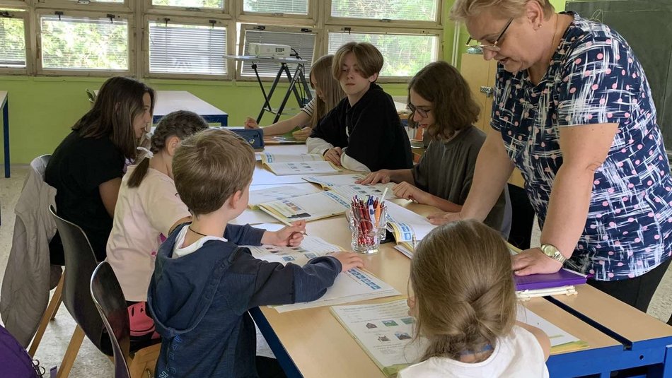 Sechs Kinder und eine Lehrerin stehen und sitzen an einem Tisch und lernen.