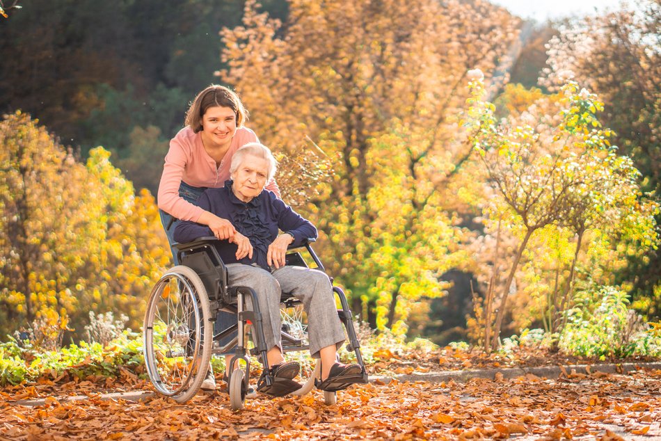 Bild zeigt eine ältere Frau die im Rollstuhl sitzt mit einer Frau in Begleitung, sie stehen vor Bäumen mit Herbstblättern