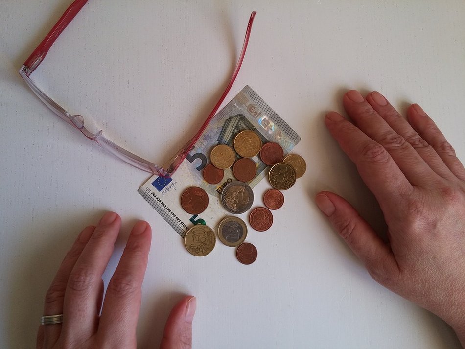 Zwei Hände liegen neben ein paar Geldscheinen und Münzen. Daneben liegt eine Brille.