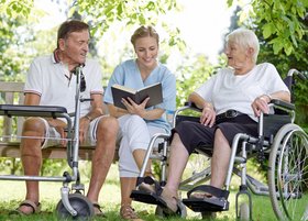 Eine Altenpflegerin sitzt im Garten zwischen zwei älteren Menschen und liest vor.