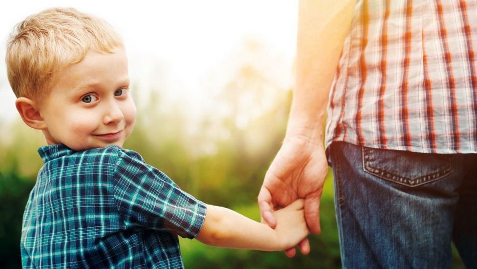 Ein kleiner blonder Junge schaut über die Schulter freundlich in die Kamera und hält eine erwachsene Person an der Hand.