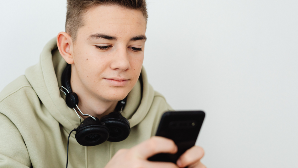 Ein Jugendlicher mit einem grauen Kapuzenpullover hat schwarze Kopfhöhrer um seine Hals hängen und ein schwarzes Smartphone in der Hand, auf das er schaut. 