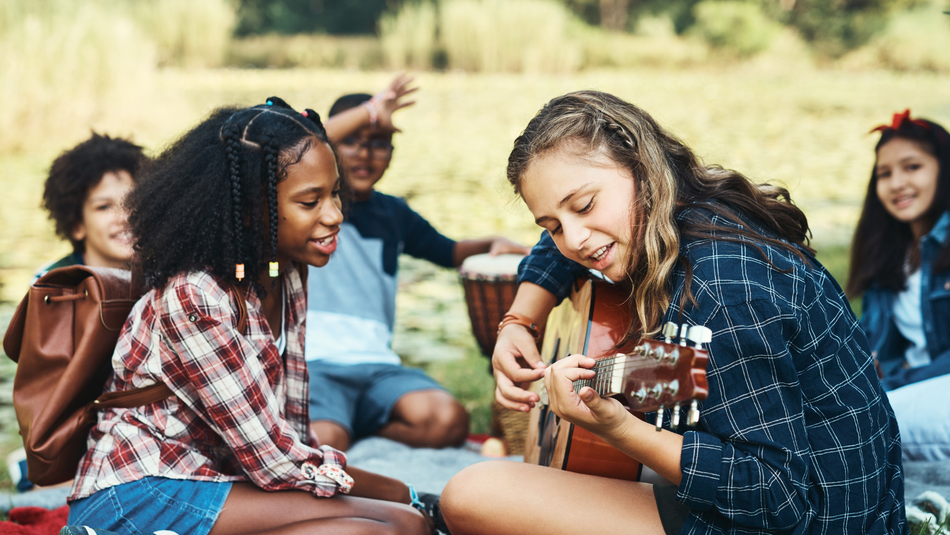 Mehrere Jugendliche sitzen auf einer Wiese. Ein Mädchen im Vordergrund spielt Gitarre.