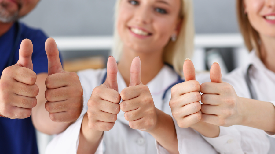Mitarbeitende im Gesundheitswesen halten mit beiden Händen ihre Daumen nach oben und lachen in die Kamera.