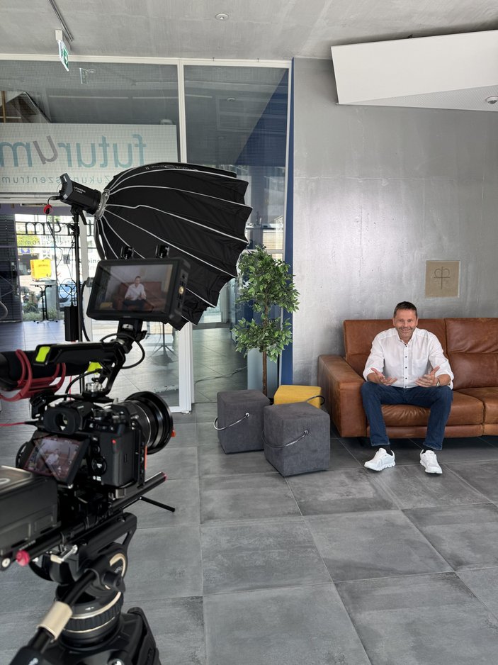 Finanzvorstand Dr. Bachert sitzt auf einem braunen Sofa und wird von einer Kamera gefilmt.