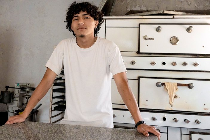 Ein junger Mann steht in einer Backstube vor einem großen Ofen, lehnt an einem Tisch und schaut in die Kamera.