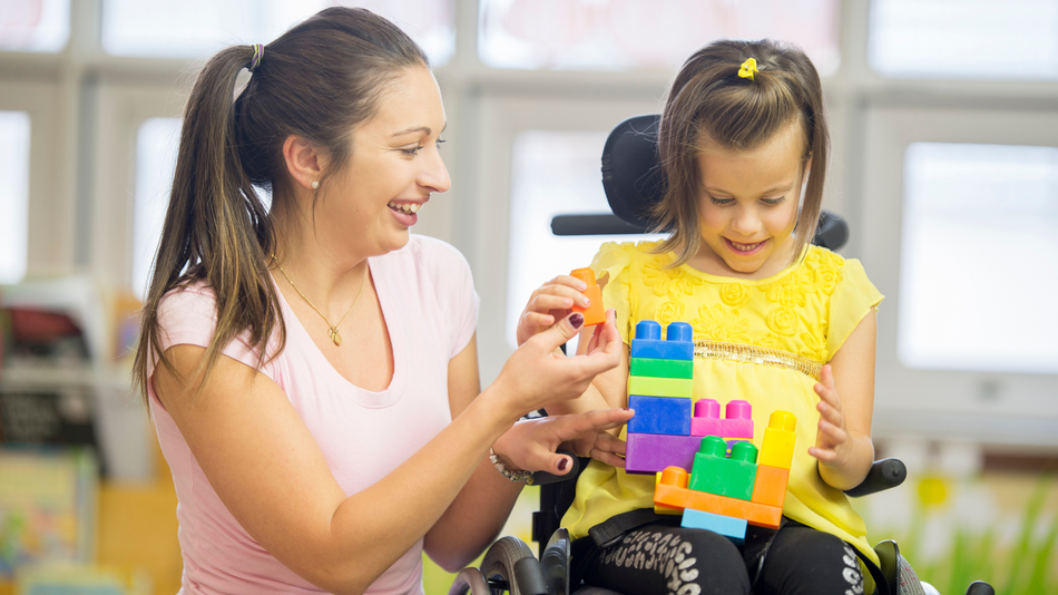 Eine Erzieherin spielt mit einem behinderten Mädchen, das im Rollstuhl sitzt. Beide lachen.