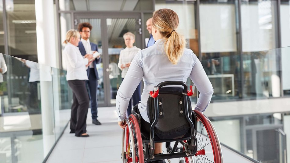Bild zeigt eine Frau die mit ihrem Rollstuhl einen gläsernen Flur entlang rollt