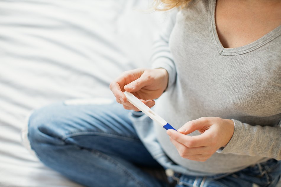 Eine Frau sitzt auf einem Bett und hält einen Schwangerschaftstest in der Hand. Ihr Kopf ist nicht zu sehen.