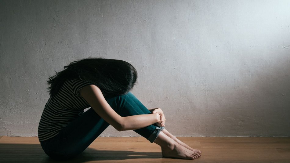 Bild zeigt eine junge Frau die mit angezogenen Beinen auf dem Boden sitzt ihr Kopf ist zwischen die Arme gesenkt