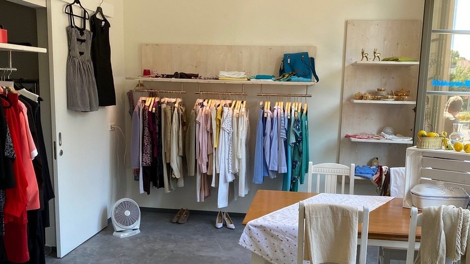 Im Vordergrund steht ein weißer Tisch mit weißen Stühlen und einer Tischdecke. Dahinter im Raum stehen Kleiderständer. An den Wänden hängen noch weitere Kleider und stehen Regale mit weiteren Kleidern.