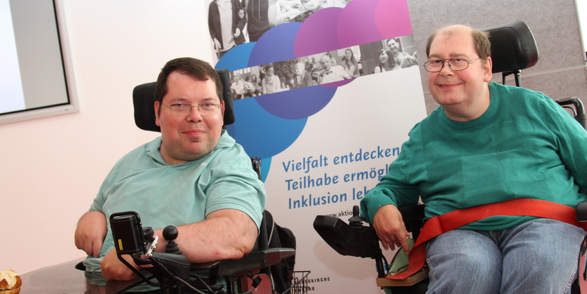 Zwei Männer mit Einschränkungen sitzen in einem Rollstuhl und lachen in die Kamera.