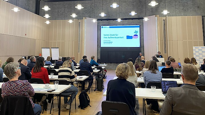 Personen sitzen an Tischen in einem großen Saal und schauen nach vorn auf eine Präsentation.