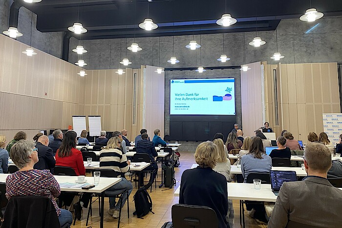 Personen sitzen an Tischen in einem großen Saal und schauen nach vorn auf eine Präsentation.