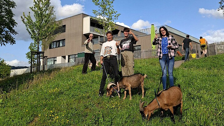 Menschen stehen mit Ziegen auf einer Wiese und strecken die Daumen nach oben