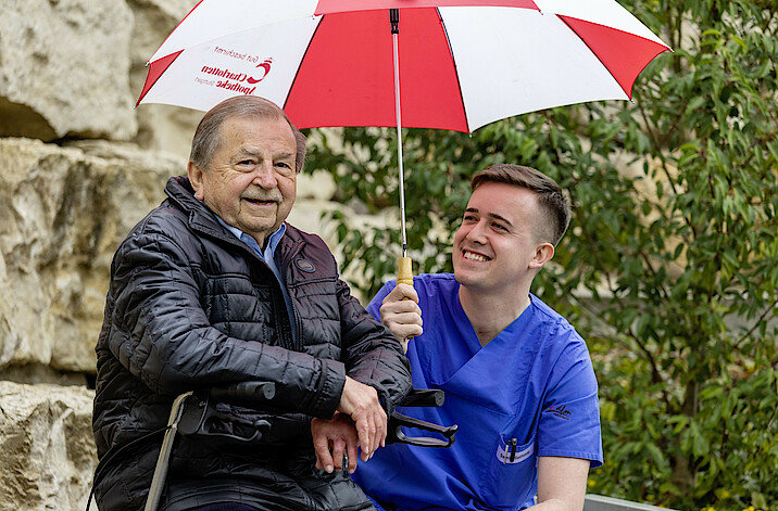 Ein junger Mann in Pflegekleidung hält lächelnd einen Regenschirm über sich und einen älteren Mann