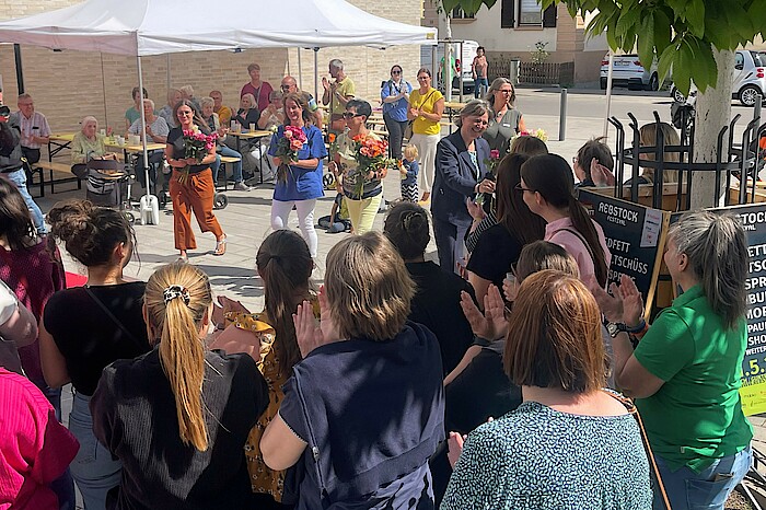 Menschen applaudieren, mehrere Frauen in der Mitte verteilen Rosen.