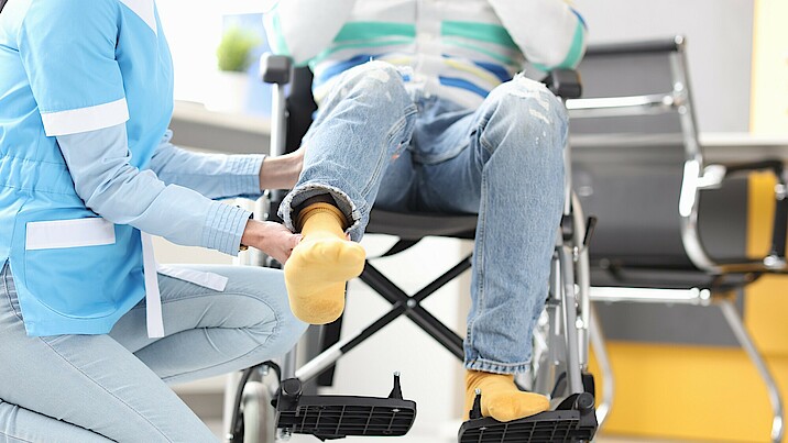 Pflegerin hebt Bein eines im Rollstuhl sitzenden Patienten. 