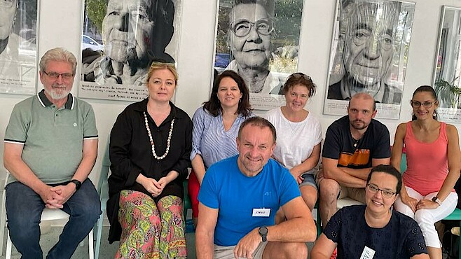 Eine Gruppe von fünf Frauen und drei Männern sitzt in einem Raum auf Stühlen und schaut lächelnd in die Kamera.