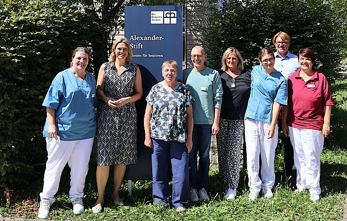 Sarah Schweizer steht neben Mitarbeitenden des Alexander-Stifts draußen vor einem Schild der Einrichtung.