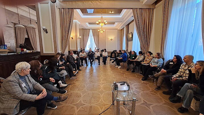 In einem großen Saal sitzen die Teilnehmenden des Workshops im Halbkreis.