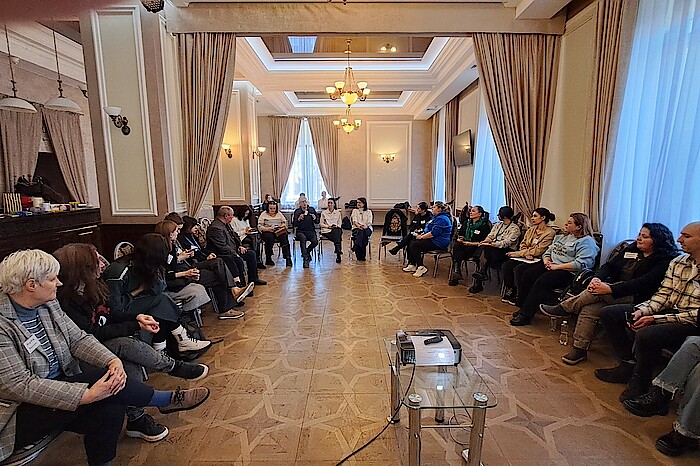 In einem großen Saal sitzen die Teilnehmenden des Workshops im Halbkreis.