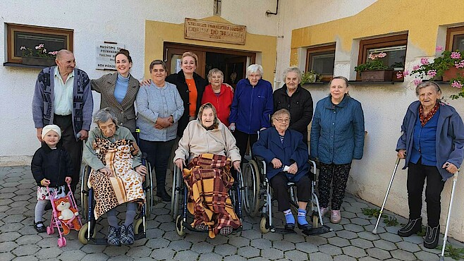 Eine Gruppe von Senioren, Seniorinnen, Pflegerinnen und einem Kind stehen vor einem Haus und lachen in die Kamera.