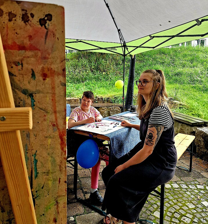 Eine Frau und ein Kind sitzen draußen an einem Tisch mit Malutensilien 